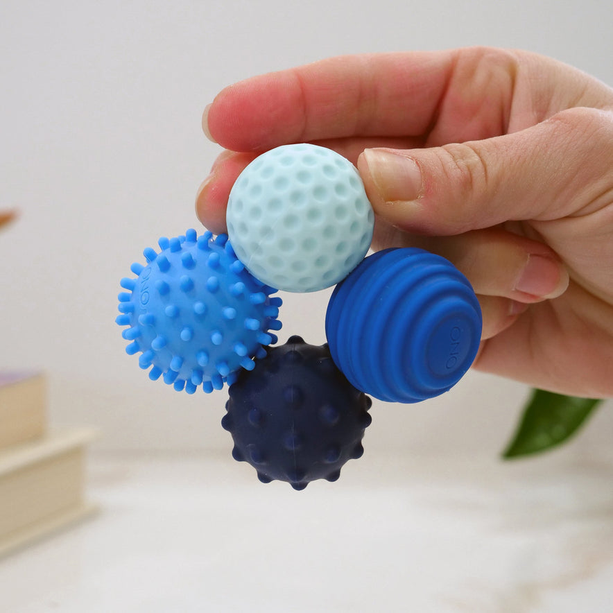 A hand holds four Signets—textured sensory balls in shades of blue—each providing a unique fidgeting experience to boost focus, set against a soft, blurred background with books and a green leaf.