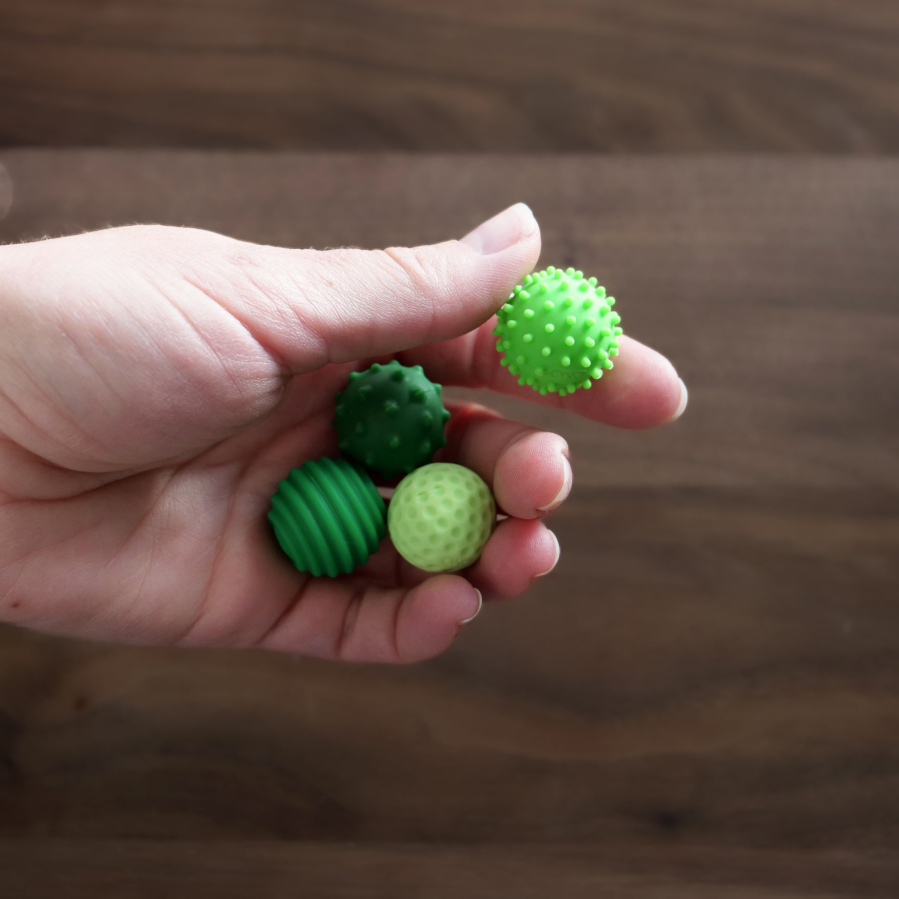 A hand holds four ONO Sigbits—small green sensory balls with smooth, spiky, and ridged surfaces—ideal as fidget toys for sensory stimulation—set against a wooden background.