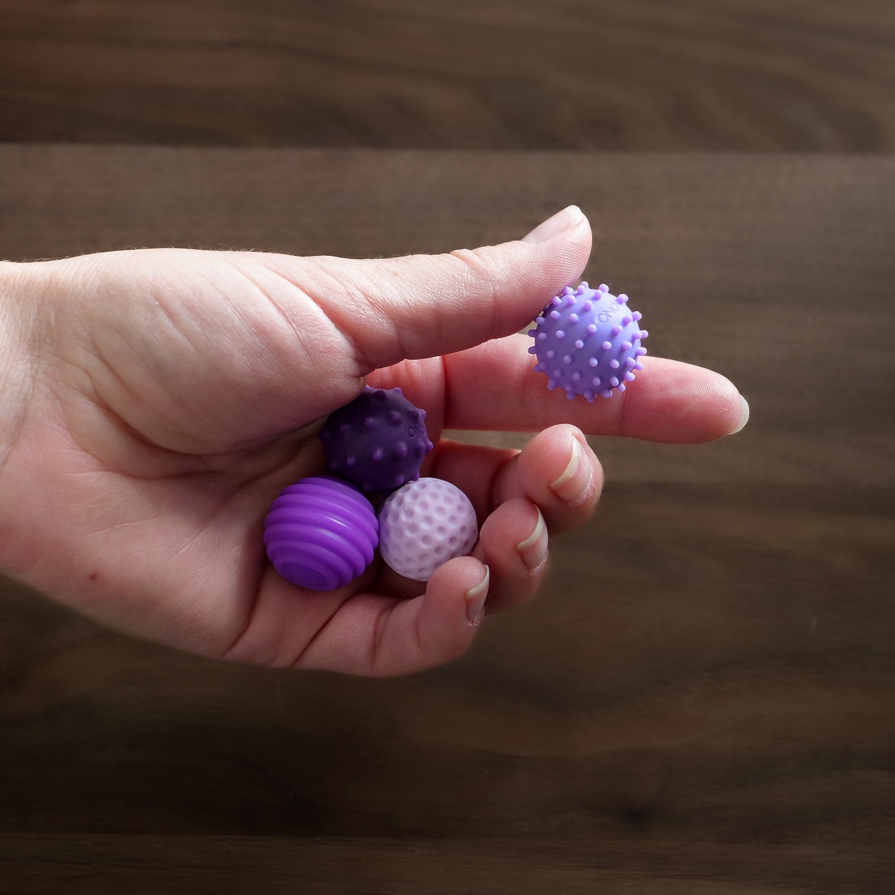 A hand holds four ONO Sigbits sensory balls in shades of purple, each with a unique textured surface—perfect as a fidget toy for sensory stimulation—against a wooden background.