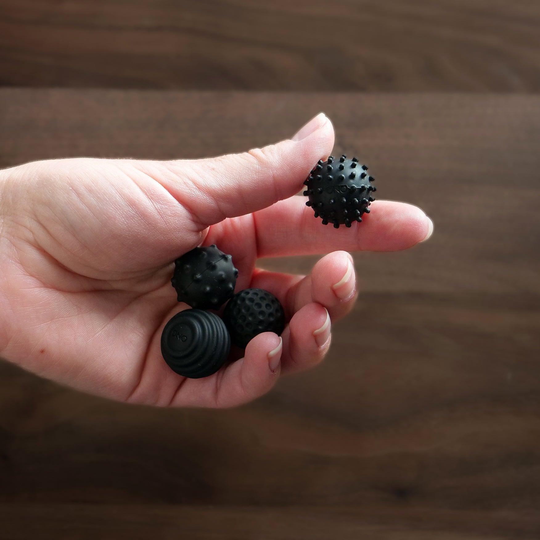 A hand holds four ONO Sigbits—small black fidget balls, each with a distinct textured surface for sensory stimulation, set against a wooden background.