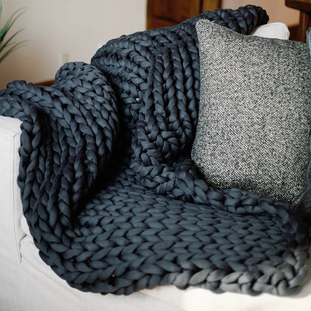 A Knit Weighted Blanket in dark blue is draped over a light-colored sofa beside a textured gray and white pillow.
