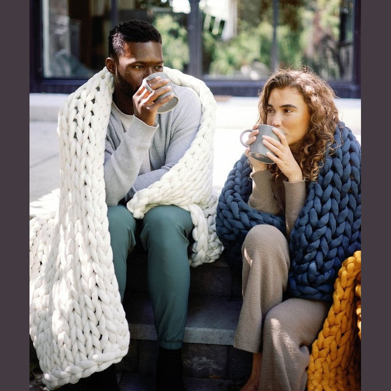 A man and a woman sit on outdoor steps, wrapped in Knit Weighted Blankets and sipping from mugs. They look relaxed and cozy, with greenery and large windows in the background.