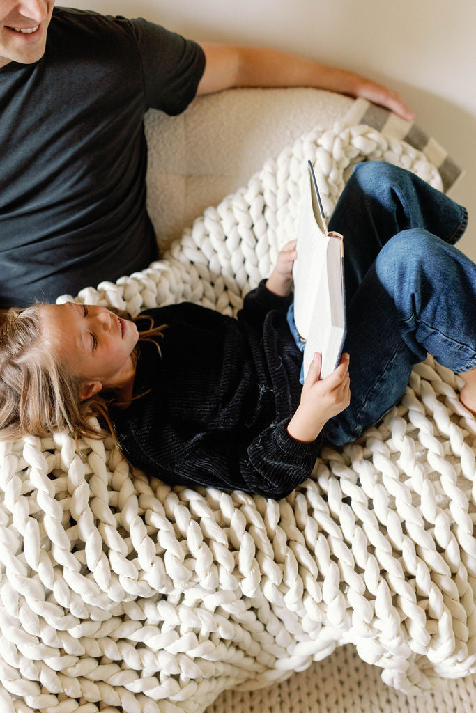 A child relaxes on the Knit Weighted Blanket, resting their head on an adult’s lap while reading, dressed in a black sweater and jeans; only part of the adult in a dark shirt is visible.