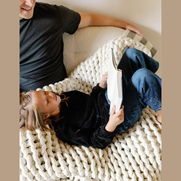 A child relaxes on the Knit Weighted Blanket, resting their head on an adult’s lap while reading, dressed in a black sweater and jeans; only part of the adult in a dark shirt is visible.