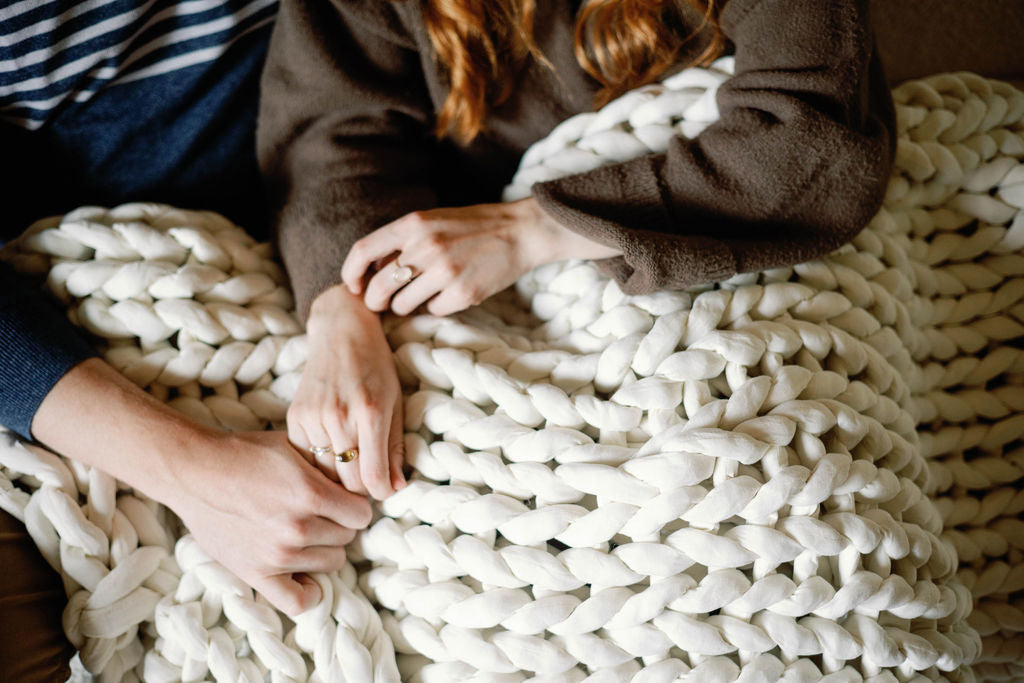 Two people sit close together, holding hands under the Knit Weighted Blanket. Only their hands, arms, and part of their torsos are visible, creating a cozy and intimate atmosphere.