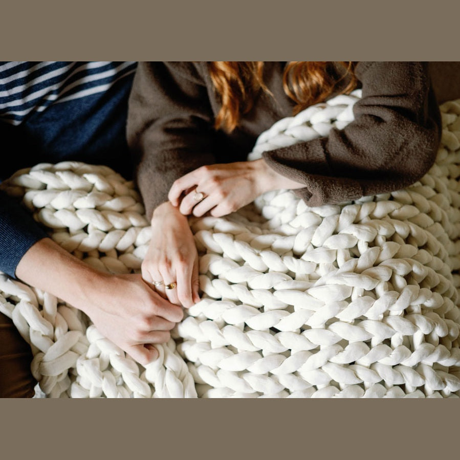 Two people sit close together, holding hands under the Knit Weighted Blanket. Only their hands, arms, and part of their torsos are visible, creating a cozy and intimate atmosphere.