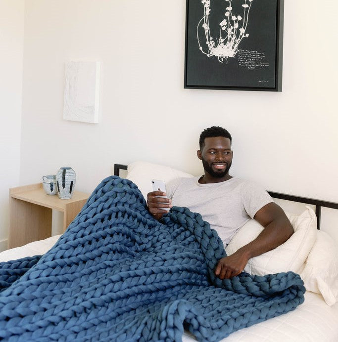 A man sits on a bed, smiling and holding a phone, wrapped in a super soft Knit Weighted Blanket. Modern art hangs on the white wall behind him, and a small table with decor is beside the bed.