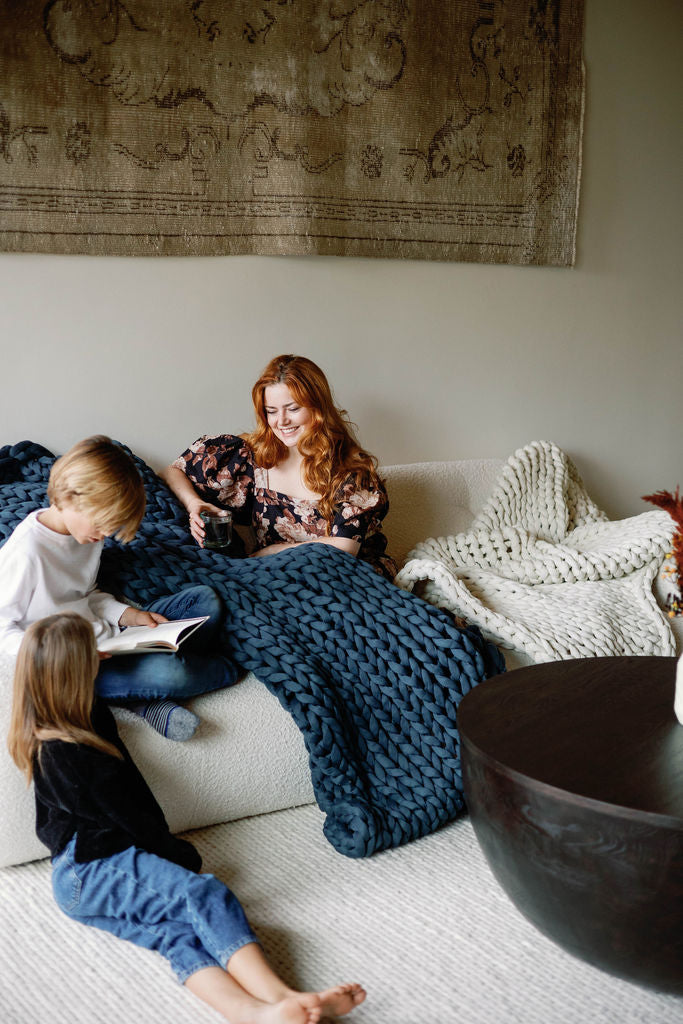 A red-haired woman smiles on a sofa with a Knit Weighted Blanket, holding a mug. Two children sit nearby—one reading—in a cozy living room with warm décor.