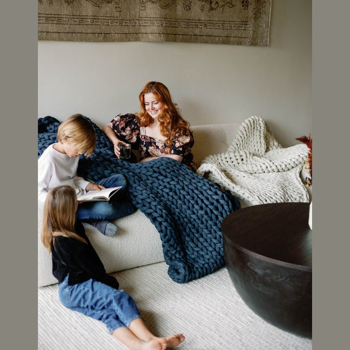 A red-haired woman smiles on a sofa with a Knit Weighted Blanket, holding a mug. Two children sit nearby—one reading—in a cozy living room with warm décor.
