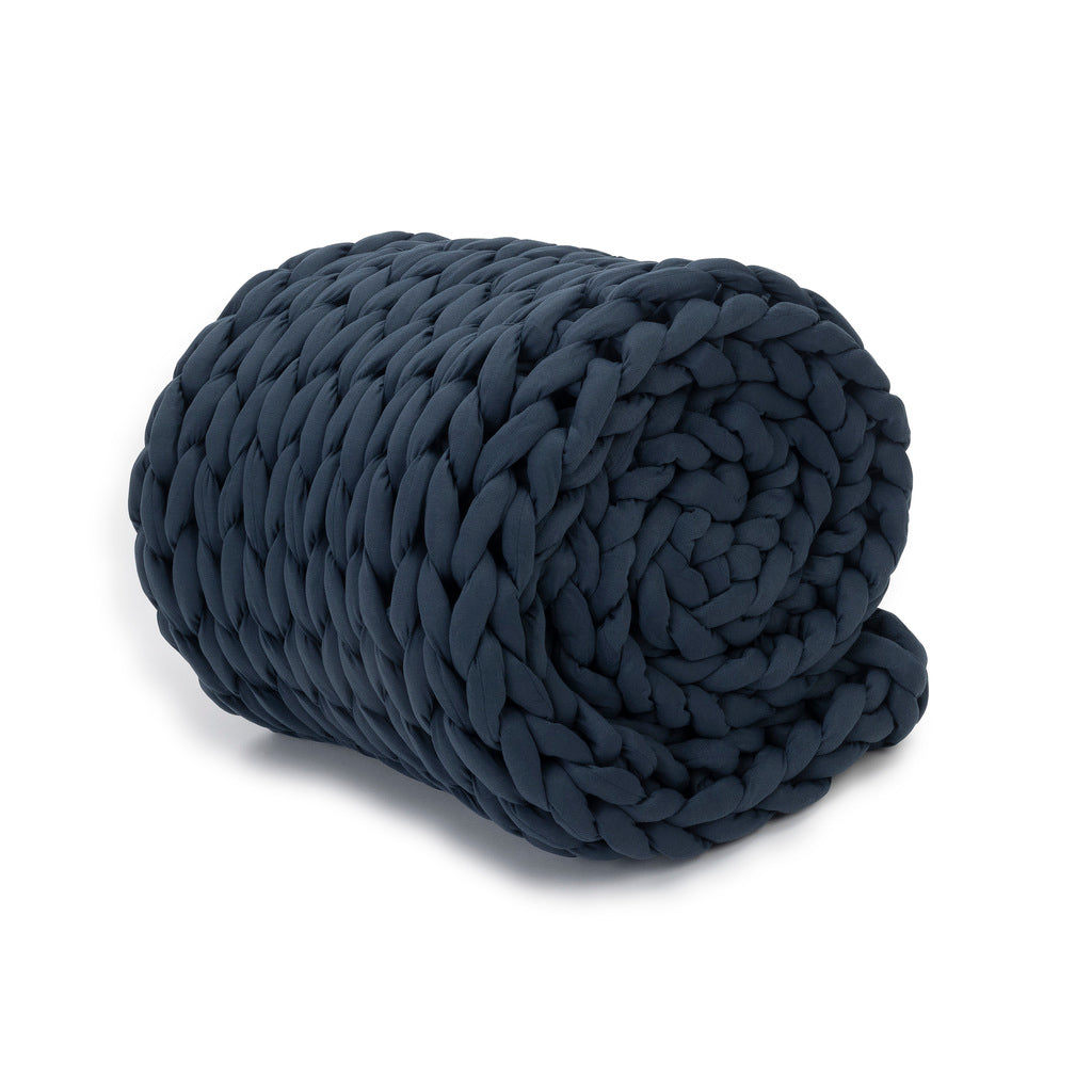 The Knit Weighted Blanket, shown tightly rolled in dark blue, highlights its thick braided texture and cozy feel.