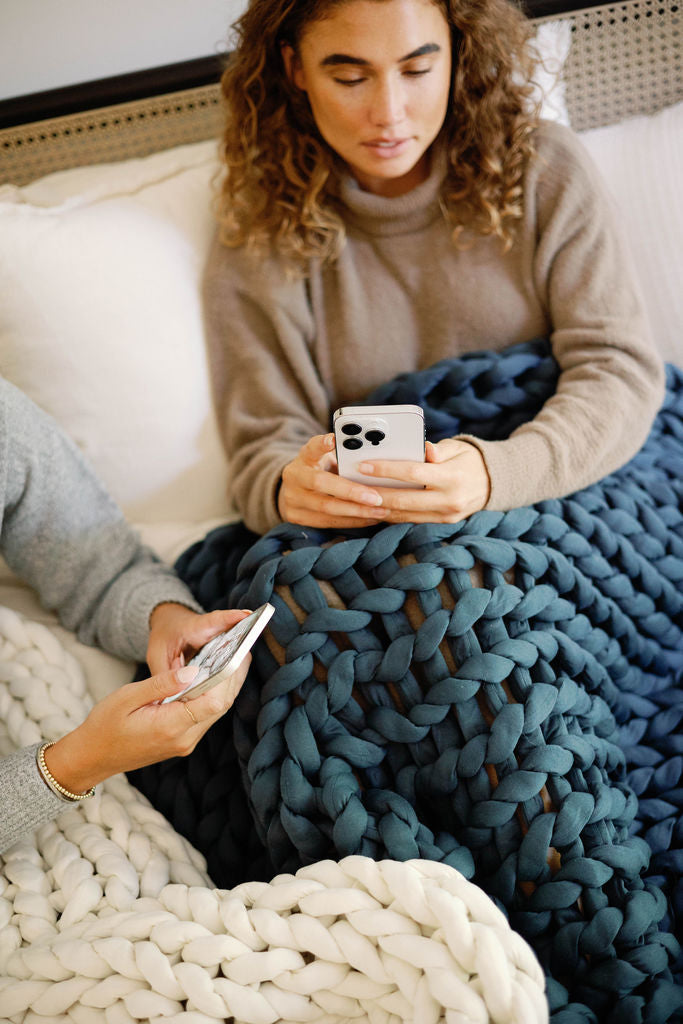 Two people sit on a bed covered with chunky knit blankets, including the Knit Weighted Blanket in blue and white. Each holds a smartphone—one wears a tan sweater, both focused on their screens.