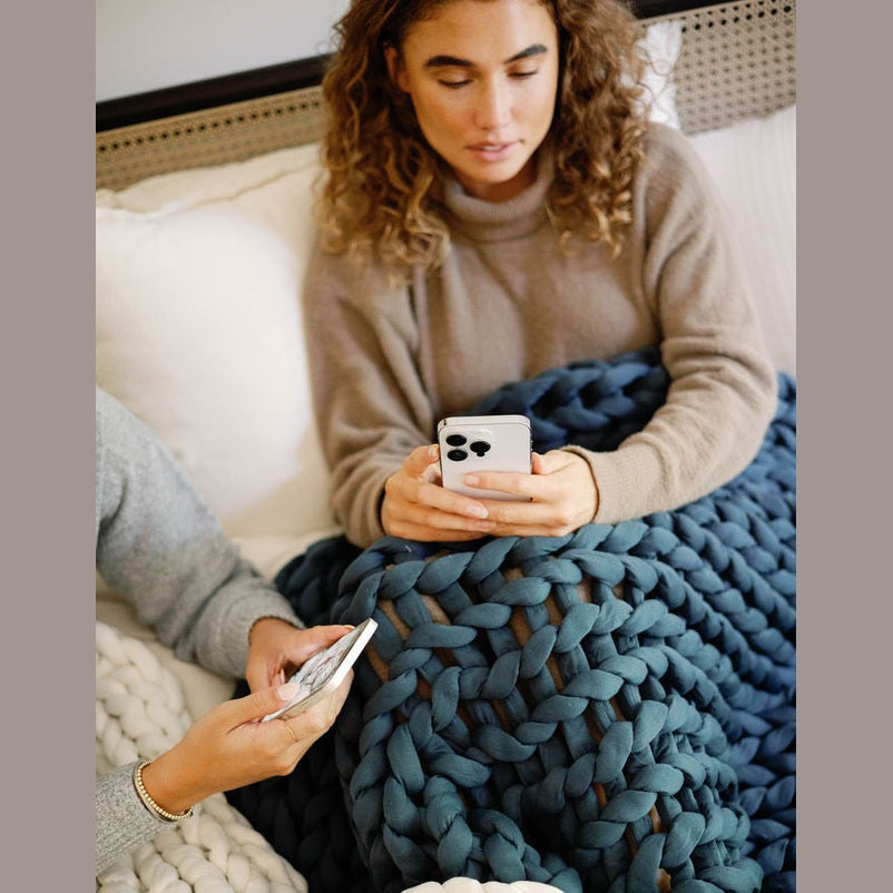 Two people sit on a bed covered with chunky knit blankets, including the Knit Weighted Blanket in blue and white. Each holds a smartphone—one wears a tan sweater, both focused on their screens.