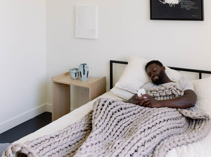A man relaxes in bed under the Knit Weighted Blanket, browsing his phone. Beside him, a small wooden table holds two ceramic mugs and a vase in a simply decorated room.