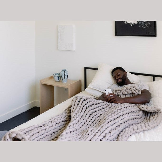 A man relaxes in bed under the Knit Weighted Blanket, browsing his phone. Beside him, a small wooden table holds two ceramic mugs and a vase in a simply decorated room.