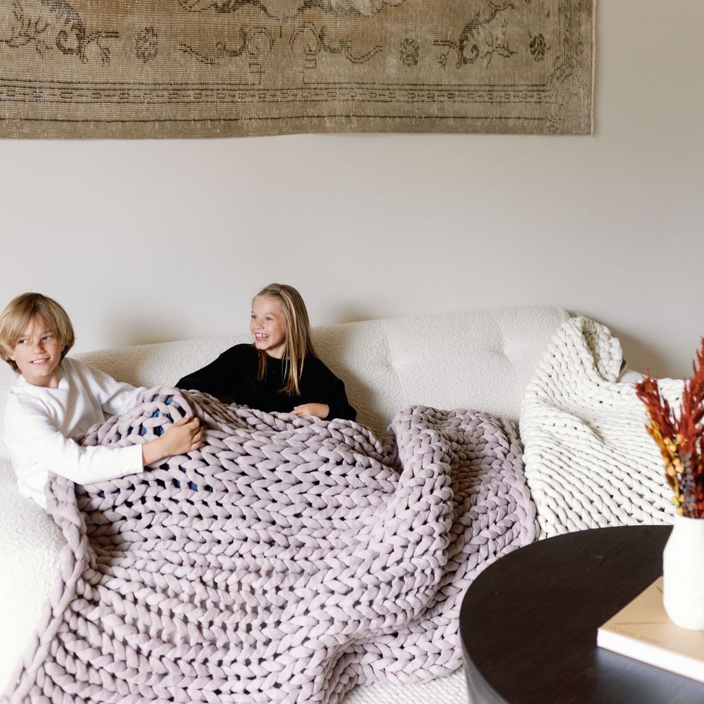 Two children smile on a couch, wrapped in a Knit Weighted Blanket. Behind them hangs a woven tapestry, while a nearby table displays a vase of dried flowers.