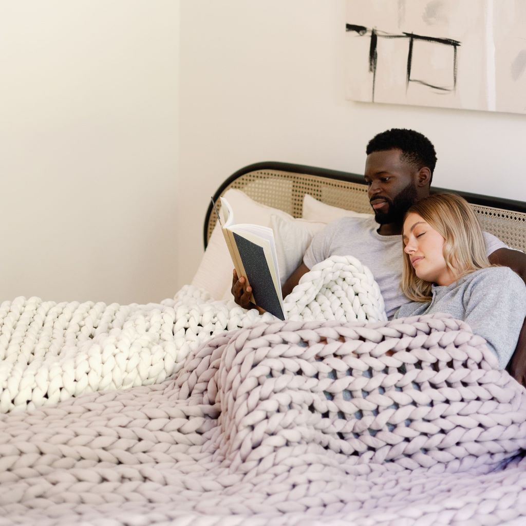 A man and woman relax in bed under a Knit Weighted Blanket; he reads a book while she rests her head on his shoulder, both looking content.