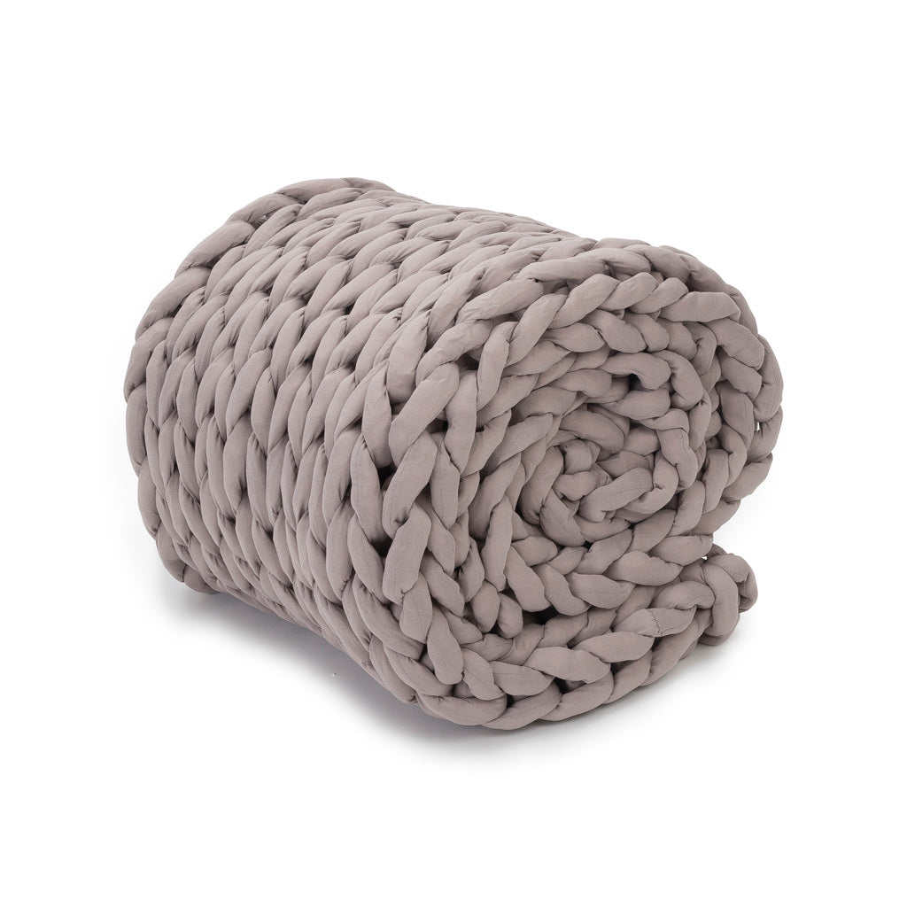 The Knit Weighted Blanket, in chunky light gray, is rolled up to show off its thick braided texture and ultra-soft, cozy look against a white background.