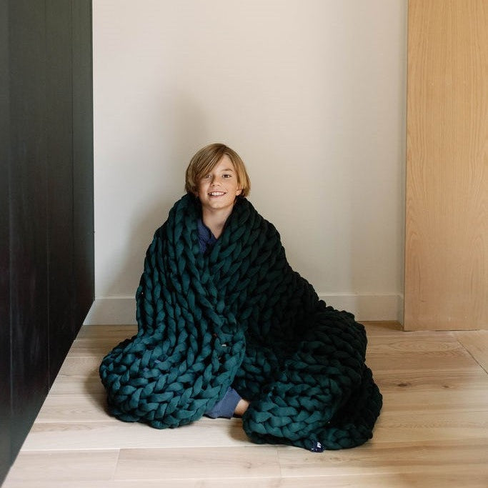 A smiling child sits on a light wooden floor, wrapped in a dark green Knit Weighted Blanket with chunky knit, against a light-colored wall featuring black and wood paneling.