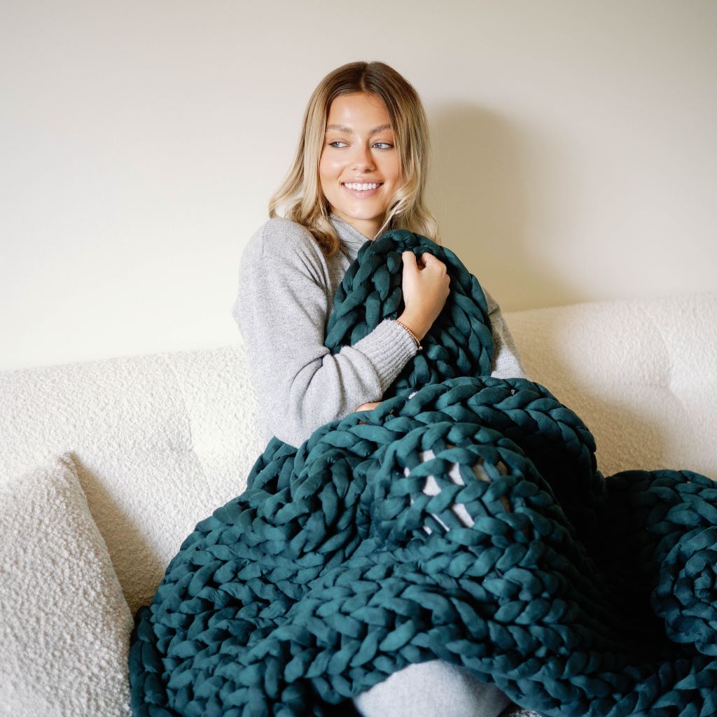 A woman, cozy and relaxed in a light gray sweater, smiles while wrapped in the Knit Weighted Blanket—a chunky dark green braided throw—sitting on a white sofa.