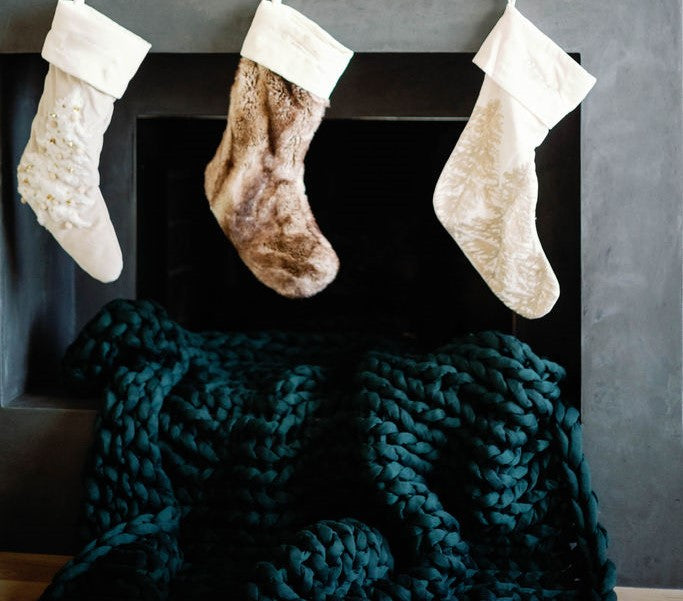 Three Christmas stockings hang above a dark fireplace, with a Knit Weighted Blanket in chunky, dark green knit draped super softly in front of the hearth.