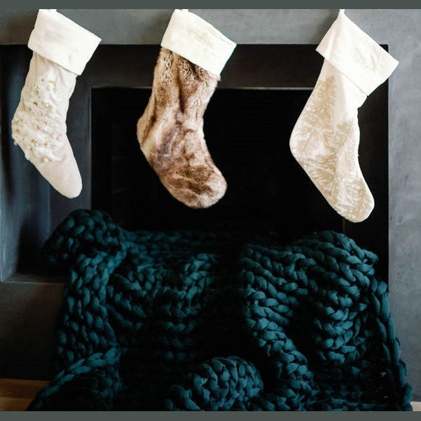 Three Christmas stockings hang above a dark fireplace, with a Knit Weighted Blanket in chunky, dark green knit draped super softly in front of the hearth.