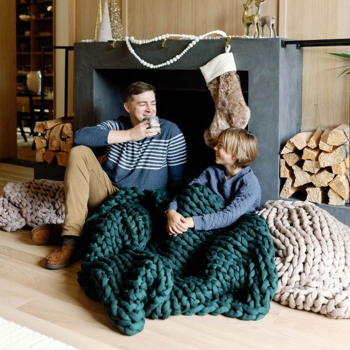 Two people sit on the floor by a fireplace, wrapped in a Knit Weighted Blanket. One smiles while sipping from a mug. Firewood and a furry stocking add warmth to the cozy scene.
