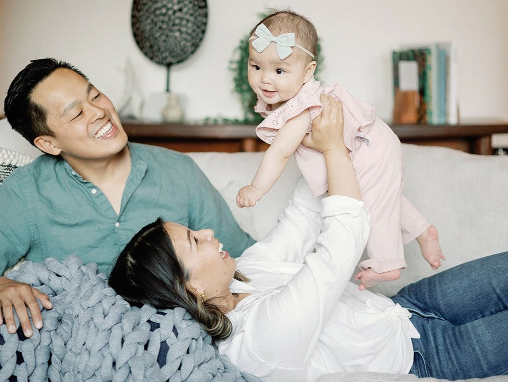 A smiling woman lifts a happy baby girl in a pink outfit and blue bow headband while they relax under the super soft Knit Weighted Blanket; a man in a green shirt sits beside them on the couch.