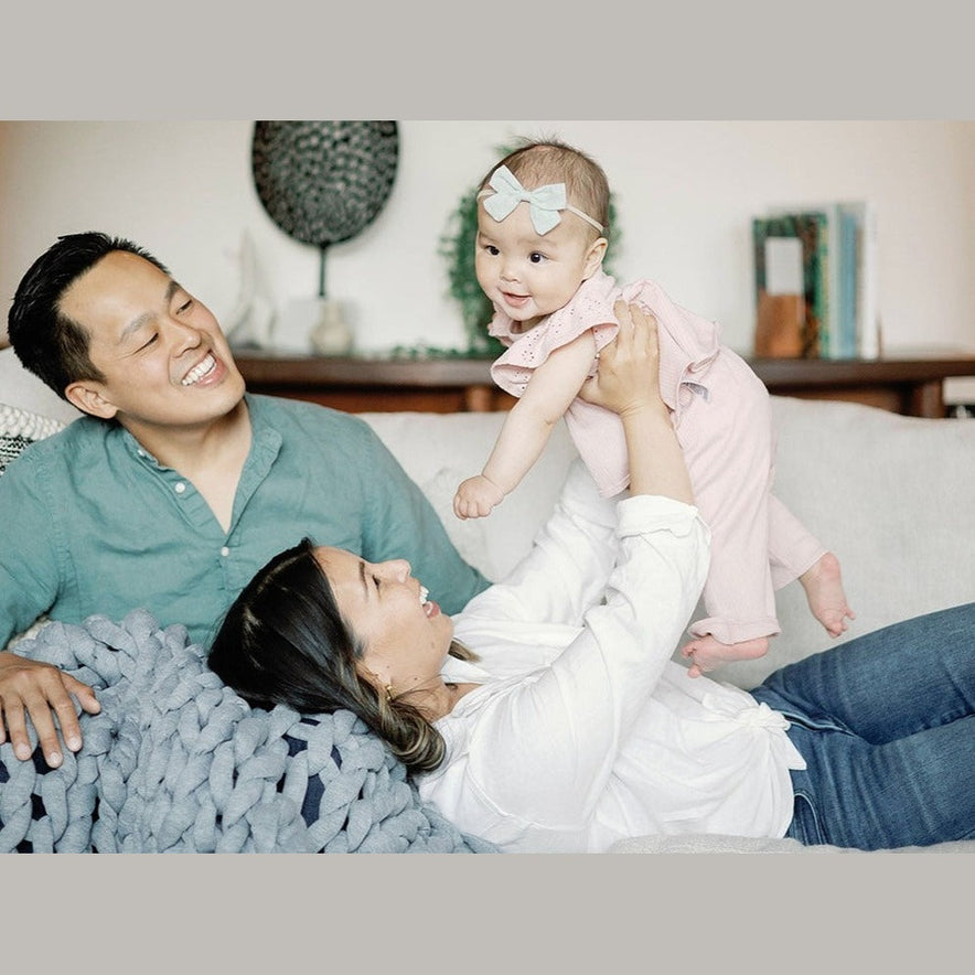 A smiling woman lifts a happy baby girl in a pink outfit and blue bow headband while they relax under the super soft Knit Weighted Blanket; a man in a green shirt sits beside them on the couch.
