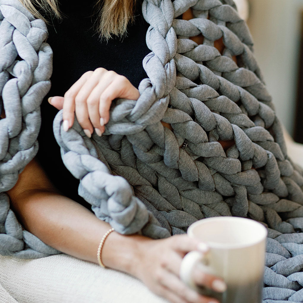 A person with manicured nails holds the Knit Weighted Blanket in chunky gray knit, wearing a black top and bracelet, and holding a white mug, creating a cozy and relaxed atmosphere.