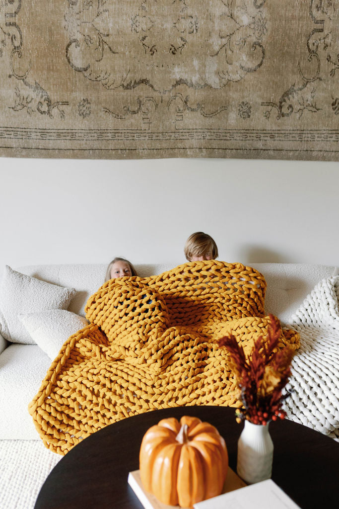 Two children are cuddled on a white sofa under the Knit Weighted Blanket. A table with a pumpkin and dried flowers sits in front, while a patterned rug decorates the wall behind them.