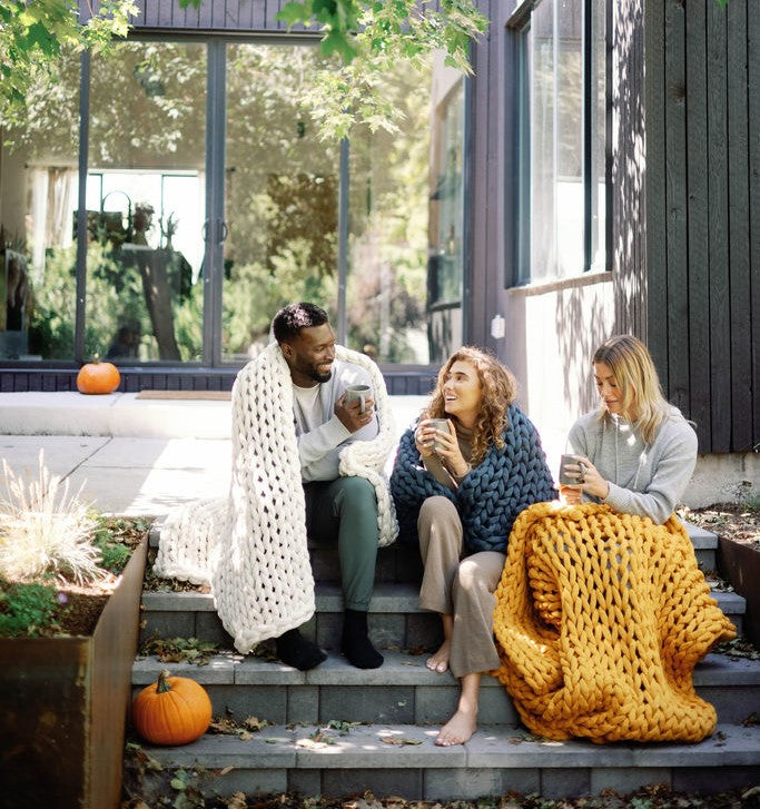 Three people sit on outdoor steps wrapped in Knit Weighted Blankets, smiling and sipping hot drinks. Pumpkins are around them, with a modern house featuring large windows in the background.