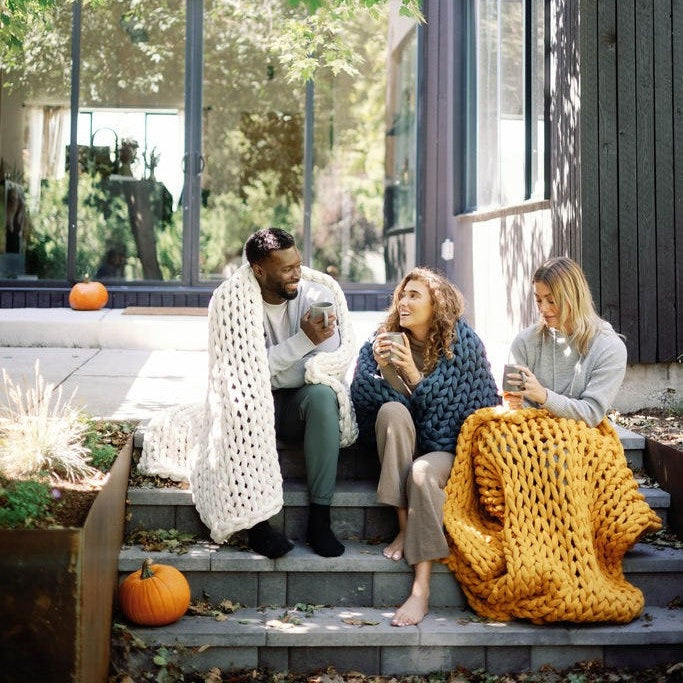 Three people sit on outdoor steps wrapped in Knit Weighted Blankets, smiling and sipping hot drinks. Pumpkins are around them, with a modern house featuring large windows in the background.