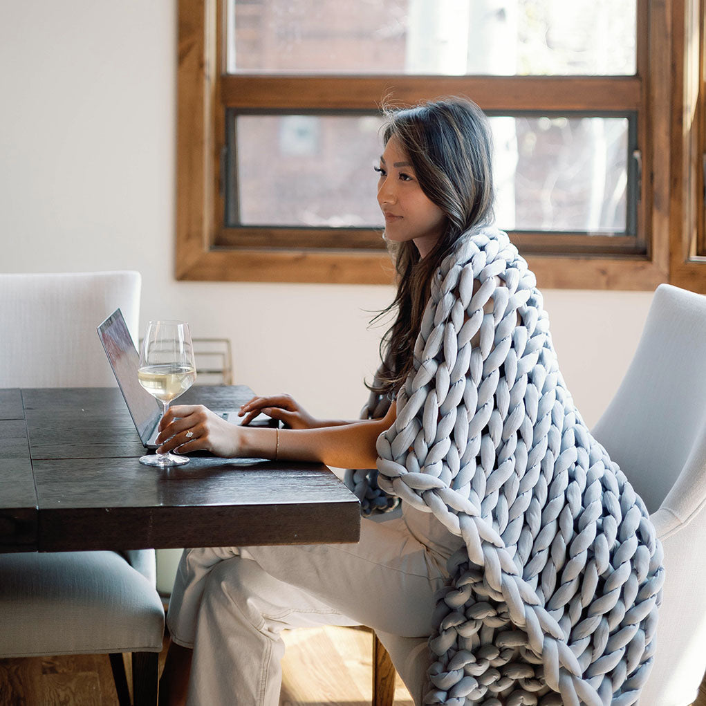 Wrapped in a Knit Weighted Blanket, a person sits at a table using a laptop and holding a glass of white wine in a bright room with modern furniture and a large window.