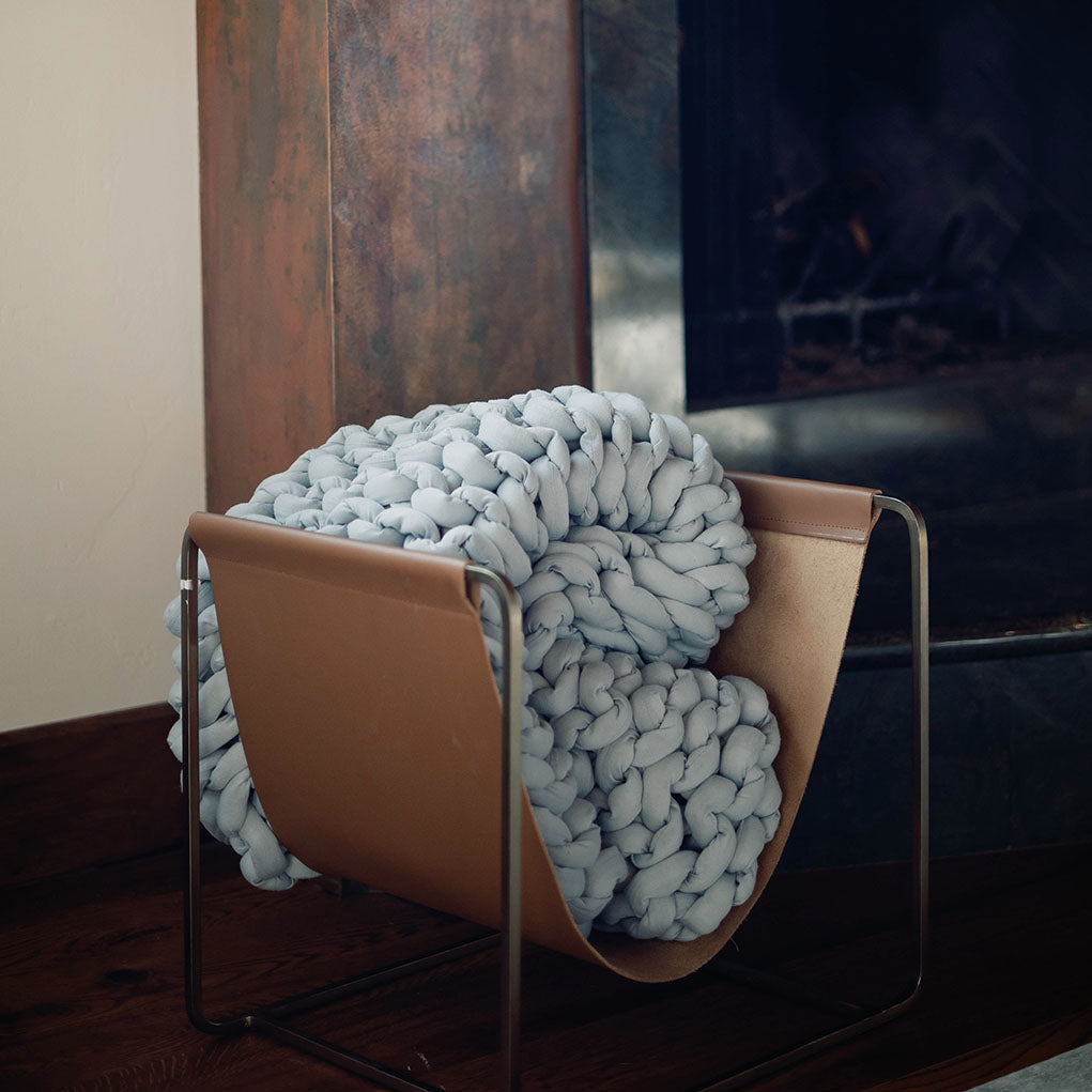 A thick, light blue Knit Weighted Blanket is folded and placed in a modern tan and metal storage holder next to a fireplace with a dark metal frame.
