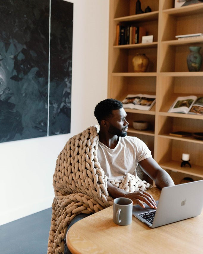 A man wrapped in a Knit Weighted Blanket sits at a table with a laptop and mug, looking to the side. Shelves with books and decor are behind him, along with a large abstract painting on the wall.