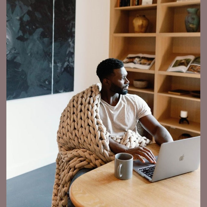 A man wrapped in a Knit Weighted Blanket sits at a table with a laptop and mug, looking to the side. Shelves with books and decor are behind him, along with a large abstract painting on the wall.