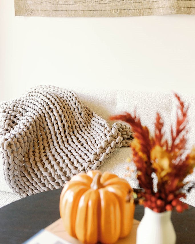 A cozy scene with a Knit Weighted Blanket draped over a white sofa. In the foreground, a ceramic pumpkin and a vase of autumn-hued dried flowers rest on a table.