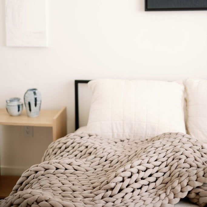 A neatly made bed with two white pillows and a beige Knit Weighted Blanket. Next to the bed, a small wooden nightstand holds two ceramic vases. The room has minimal, modern decor and light-colored walls.