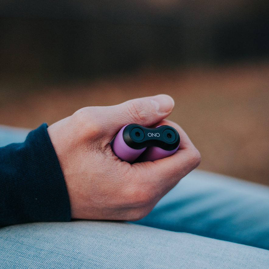 A hand holds a small, purple and black ONO Roller Soft Touch Plastic fidget toy with the thumb on top—ideal for stress relief—against a softly blurred background.