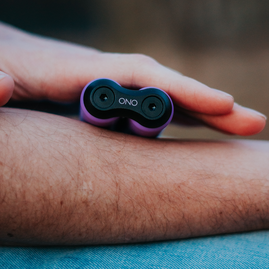 A close-up of a hand holding the ONO Roller Soft Touch Plastic, a small purple silent fidget toy, against their forearm with a blurred background.