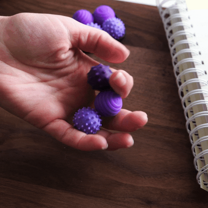 A hand holds ONO Sigbits—small purple textured sensory fidget balls—over a wooden table beside a spiral-bound notebook, making them an ideal fidget toy for sensory stimulation and enhanced focus.