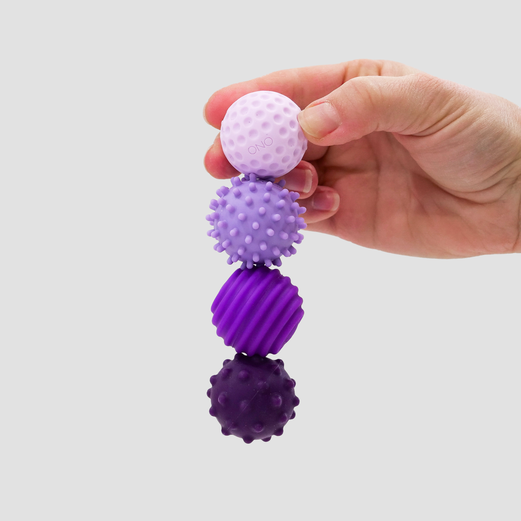 A hand holds four stacked Signets sensory balls in pink and purple hues, each featuring a unique textured surface, providing a satisfying fidget experience against a plain light gray background.