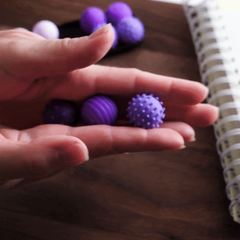 A hand holds three ONO Sigbits—small purple textured balls in different patterns—ideal as a fidget toy for sensory stimulation, shown above a wooden surface near a spiral notebook.