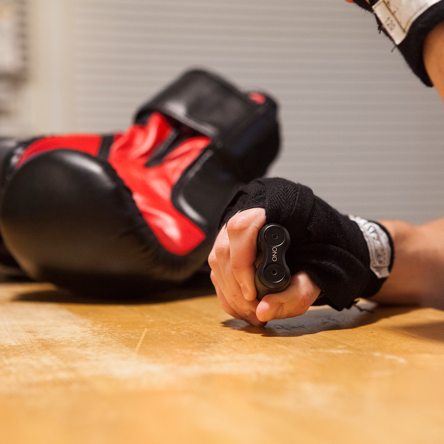 A person with wrapped hands fidgets with a pair of black ONO Roller Aluminum devices on a wooden table, while red and black boxing gloves rest in the background.