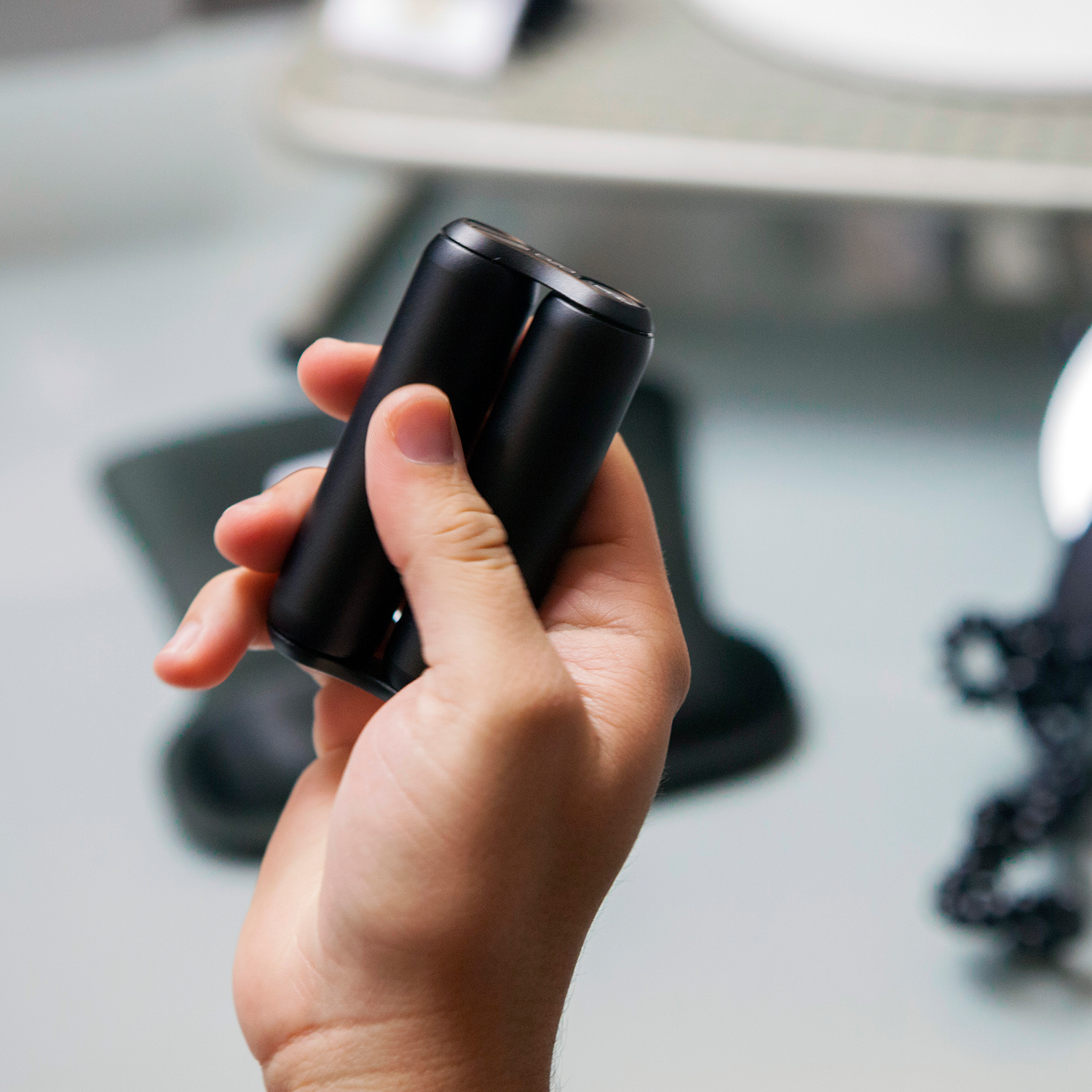 A person holds an ONO Roller Aluminum, a sleek black cylindrical device, in their hand with a blurred table background—ideal for discreet fidgeting.