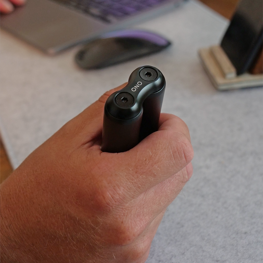 A hand holds the ONO Roller Aluminum, a sleek black cylindrical device with two top buttons, in front of a laptop, mouse, and smartphone on a desk—perfect for fidgeting and boosting focus while working.