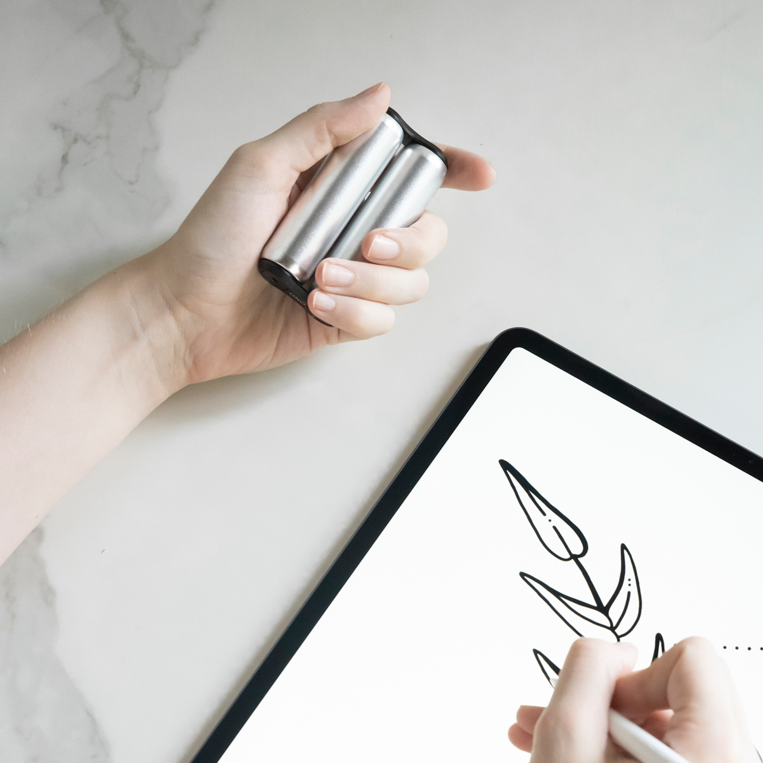 While drawing a floral design on a tablet with a stylus, a person fidgets with an ONO Roller Aluminum in their other hand, all set on a light-colored marble surface.