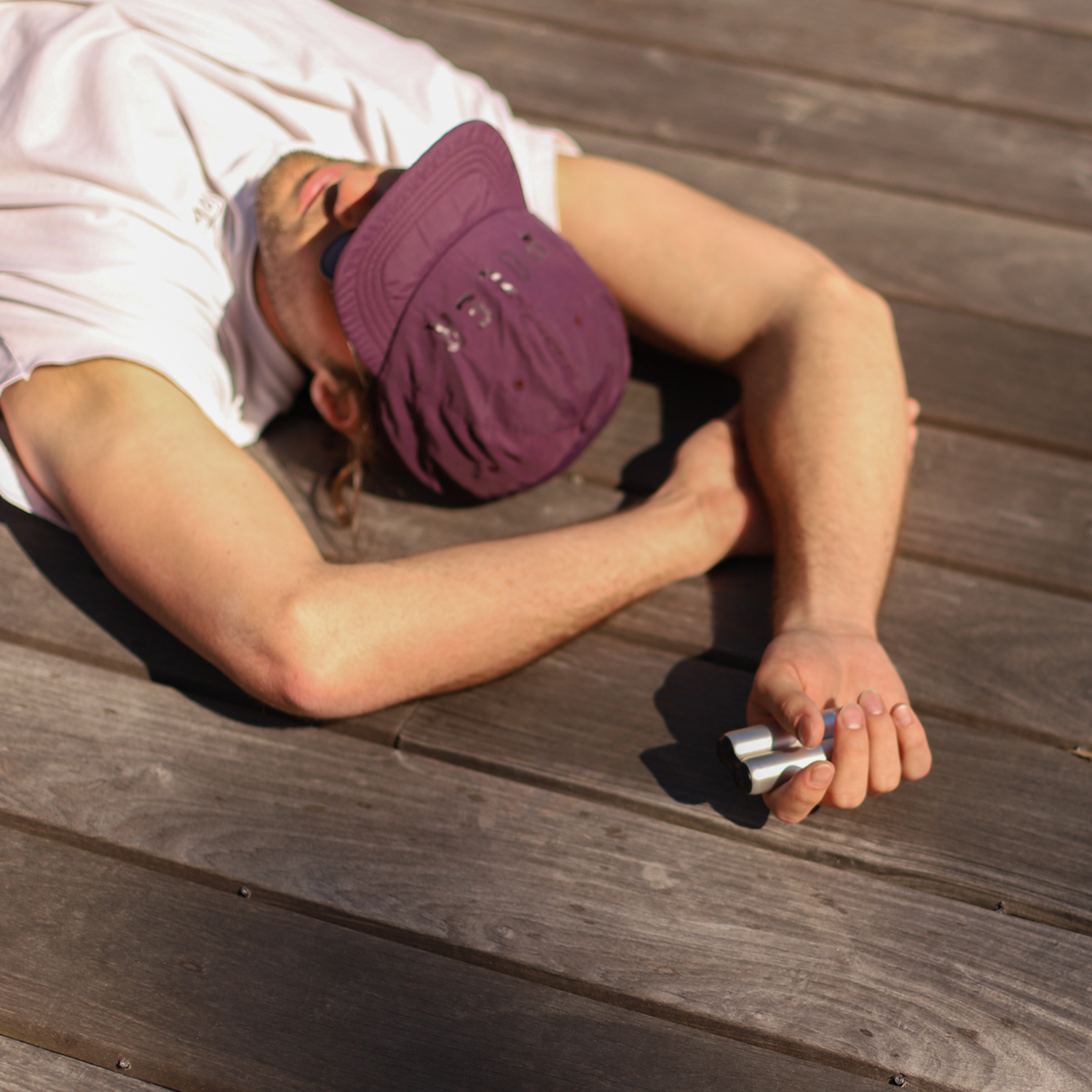 A person in a purple cap and sunglasses relaxes on a wooden deck, one arm behind their head while the other hand fidgets with an ONO Roller Aluminum.