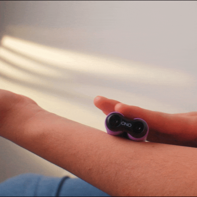 A hand rolls the ONO Roller Soft Touch Plastic, a small purple stress-relief device, over a forearm. Sunlight comes from the left, casting shadows on skin and wall as this silent roller soothes tension.