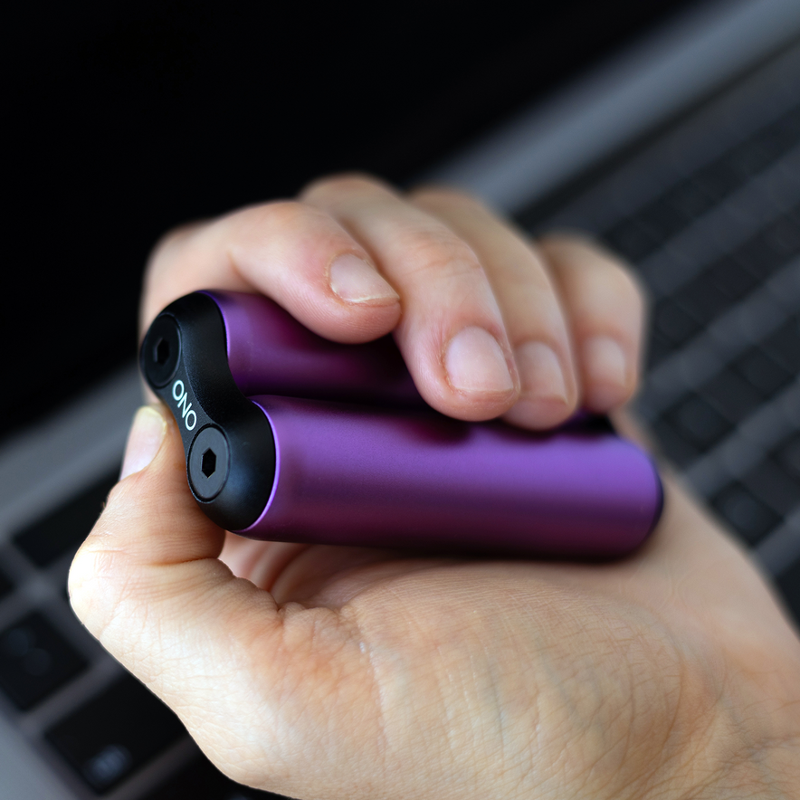 A hand holds a small, purple ONO Roller Aluminum with black ends, ideal for fidgeting, next to a laptop keyboard. ONO is printed on one end of the device.